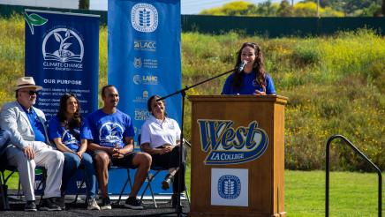 Guest speaker at podium, speaking, while fellow speakers listen on