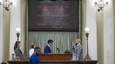 Asm. Bryan holding up hand, being sworn in by Speaker Rivas, with display screen above