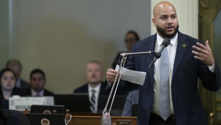 Asm. Bryan at microphone, holding paper and gesturing with one hand while speaking