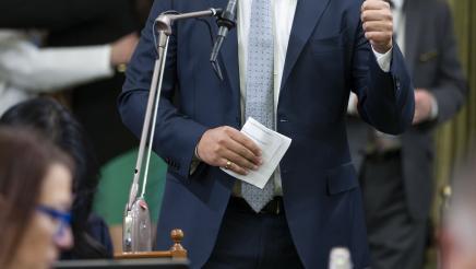 Asm. Bryan at microphone, holding paper and holding up one thumb while speaking