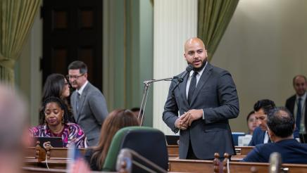 Asm. Bryan standing and speaking into microphone, with other members at left and right
