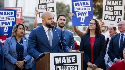 Asm. Bryan at podium, speaking, with Asm. Hart, Asm. Friedman and supporters in background holding signs