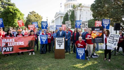 Asm. Bryan at podium, speaking, with Asm. Hart, Asm. Friedman and supporters in background holding signs