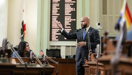 Asm. Bryan standing and speaking on the Assembly floor, gesturing with an upheld hand