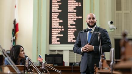 Asm. Bryan standing and speaking on the Assembly floor as a fellow member listens on