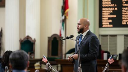 Asm. Bryan standing and speaking on the Assembly floor as other members listen on