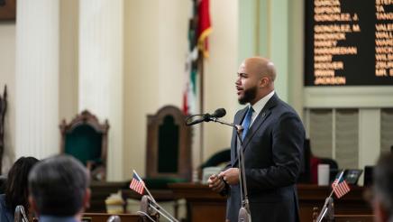 Asm. Bryan standing and speaking on the Assembly floor as other members listen on