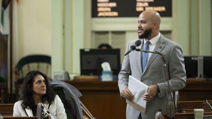 Asm. Bryan standing on the Assembly Floor and speaking into microphone as fellow member listens on
