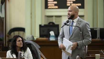 Asm. Bryan standing on the Assembly Floor and speaking into microphone as fellow member listens on