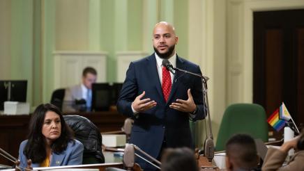 Asm. Bryan standing at microphone, speaking, as other members listen on