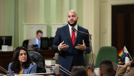Asm. Bryan standing at microphone, speaking, as other members listen on