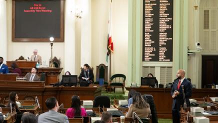 Asm. Bryan standing at microphone on Assembly Floor, speaking, as other members listen on