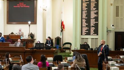 Asm. Bryan standing at microphone on Assembly Floor, speaking, as other members listen on