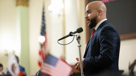 Asm. Bryan standing at microphone, speaking, with flags in foreground and background