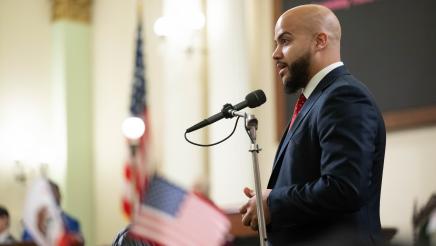 Asm. Bryan standing at microphone, speaking, with flags in foreground and background