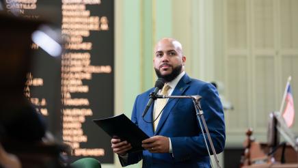 Asm. Bryan standing at microphone, speaking and holding folder