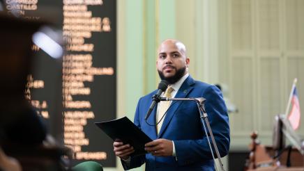 Asm. Bryan standing at microphone, speaking and holding folder