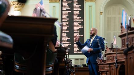 Asm. Bryan standing at microphone, speaking, gesturing with one hand and holding folder