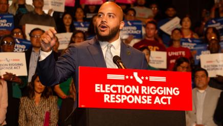 Asm. Bryan at podium, speaking and gesturing with one hand