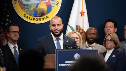 Asm. Bryan at podium, speaking, while fellow legislators listen on