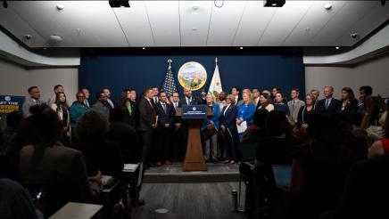 Wide view of Asm. Bryan at podium, speaking, while fellow legislators listen on