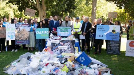Asm. Bryan at podium, speaking, with recyclables piled in foreground